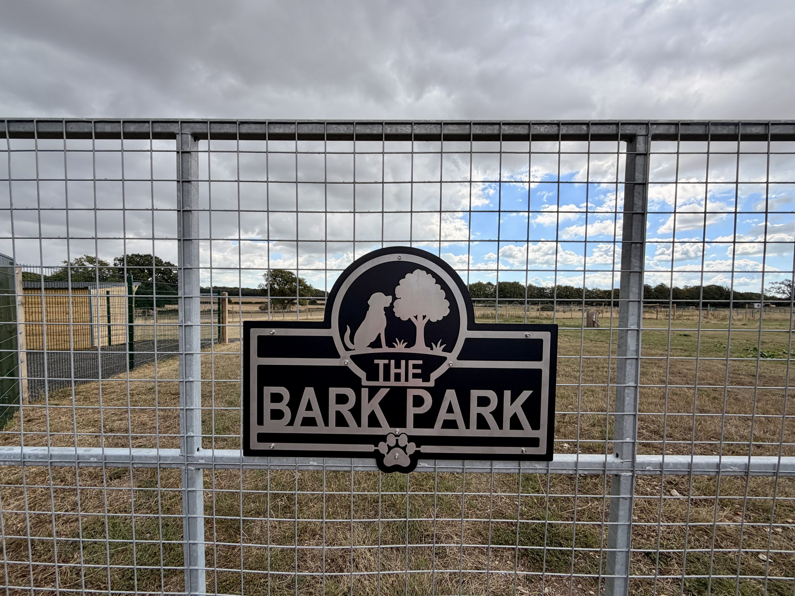 A metal gate displays a sign reading The Bark Park with dog silhouettes, a tree, and a paw print, set in front of open fields.