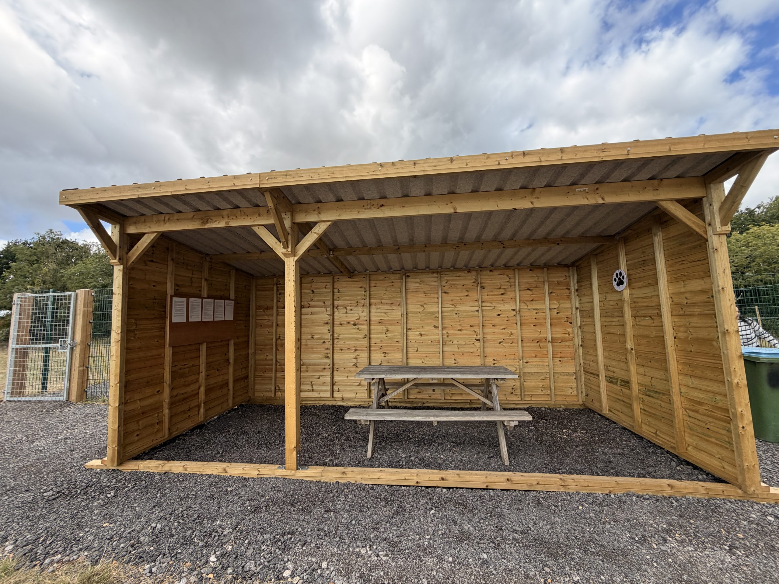A wooden shelter stands on gravel with a picnic table beneath it under a cloudy sky.